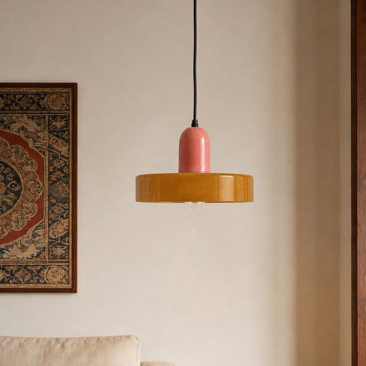 A modern pendant light with a glossy mustard yellow circular shade and a matte pink cylindrical top, hanging against a warm beige wall with a framed rug partially visible on the left.