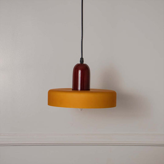 Modern pendant light with red holder and flat yellow shade hanging against neutral wall background showcasing bold contemporary lighting decor styling.