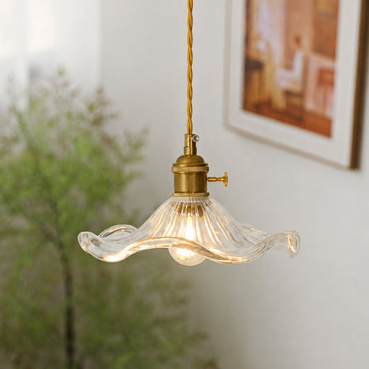 A hanging pendant light with a wavy clear glass shade and a brass-toned socket and cord, shown lit in a softly blurred indoor setting.