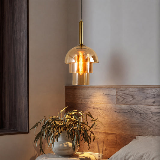 A modern pendant light with a brushed brass cylindrical neck and a clear glass dome shade, hanging above a wooden headboard in a bedroom setting with a vase of eucalyptus nearby.