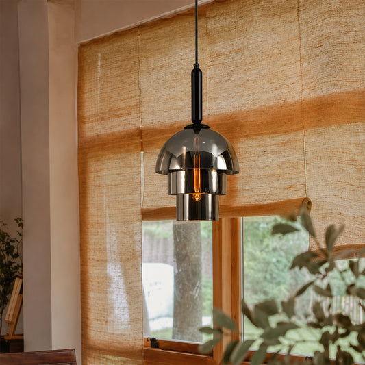 A modern pendant light with a brushed silver dome shade and black cord, hanging in front of a window with woven beige curtains and visible greenery outside.