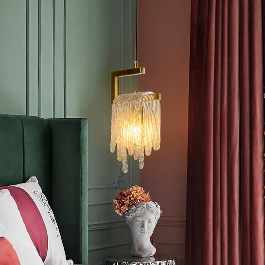 A modern wall-mounted candle-style sconce with a gold metal bracket and a textured, dripping glass shade that glows warm yellow, shown in a styled bedroom setting next to a green headboard and burgundy curtains.