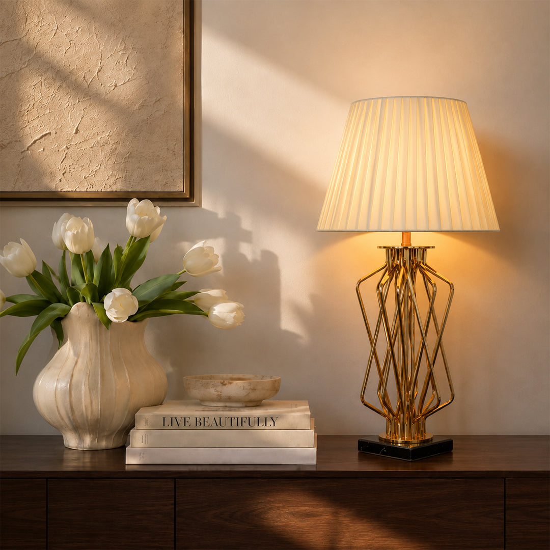 A modern table lamp with a pleated cream shade and a gold geometric wire frame base, illuminated and styled on a dark wood console table next to a vase of white tulips and stacked books.