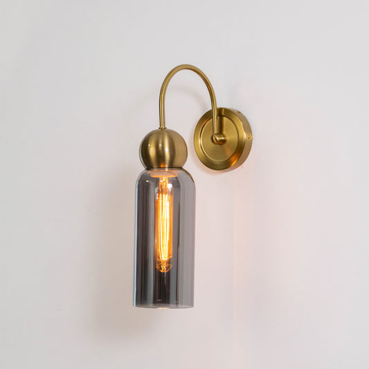 Contemporary wall light in gold metal finish with cylindrical glass shade for bedroom or living decor lighting placement white background.