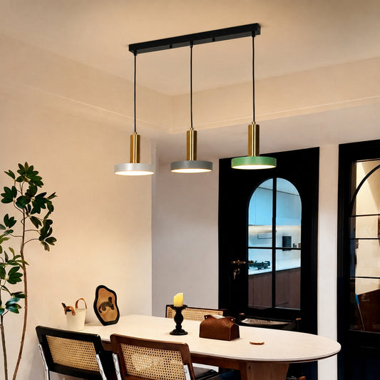 A modern three-light linear pendant chandelier hanging above a dining table in a real interior room. The fixture has a black rectangular ceiling mount, three black hanging cords, cylindrical gold-toned shades, and three round white/gray diffusers, with one light showing a green glow.