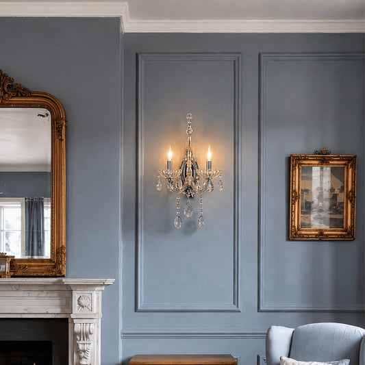 A crystal-accented wall sconce with a silver-toned metal frame and lit candle-style bulbs, mounted on a blue paneled wall in an elegant room setting.