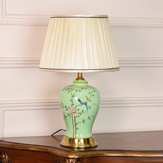 Decorative green ceramic table lamp with floral bird design brass base and fabric shade displayed on soft off white background.