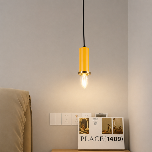 A modern cylindrical pendant light with a yellow body and gold-toned metal accents, hanging above a bedside table in a bedroom setting.