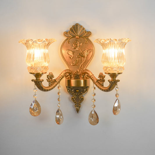 An ornate double-arm wall sconce with a gold-finished metal frame, featuring two fluted clear glass shades, hanging teardrop crystal drops, and a decorative crest plate on the backplate, shown illuminated against a plain light gray wall.
