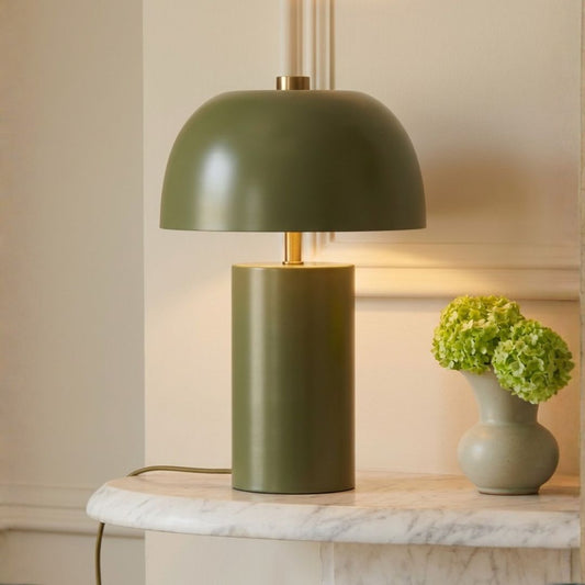 An olive green table lamp with a dome-shaped shade and cylindrical base sits on a white marble surface next to a small vase of green hydrangeas against a cream-colored wall.