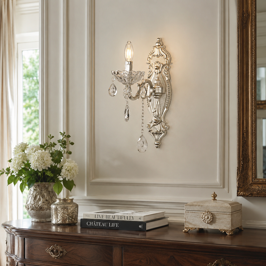 An ornate silver wall sconce with a single candle-style bulb, clear hanging crystal drops, and a decorative scrollwork design, mounted on a cream paneled wall above a dark wood console table with flowers and books.
