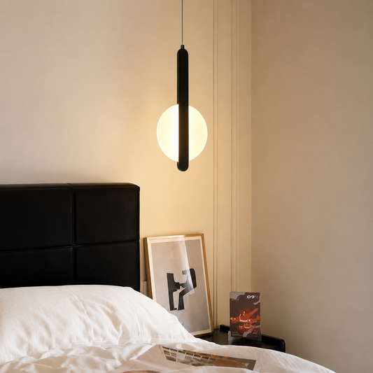 A modern black linear pendant light with a glowing white globe diffuser, hanging above a bed in a minimalist bedroom setting.