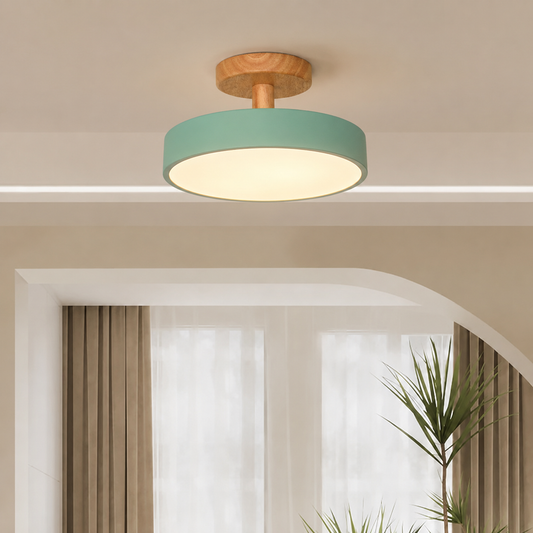 A modern semi-flush mount ceiling light with a round sage green shade, a natural wood ceiling canopy, and a warm white glowing diffuser, installed in a minimalist beige room.