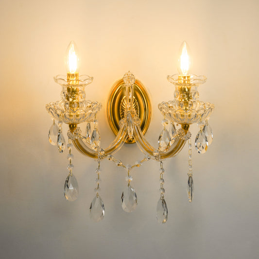 A two-light wall sconce with a polished gold-tone metal frame, clear glass crystal candle-style holders, and hanging teardrop crystal drops, shown illuminated against a plain wall.