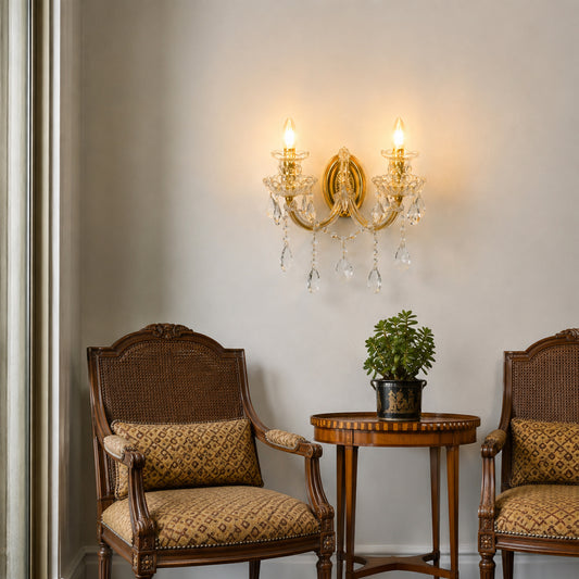 A two-light brass wall sconce with hanging crystal prisms is mounted on a beige wall above a small round table with a potted plant, flanked by two woven-back armchairs in a classic interior setting.
