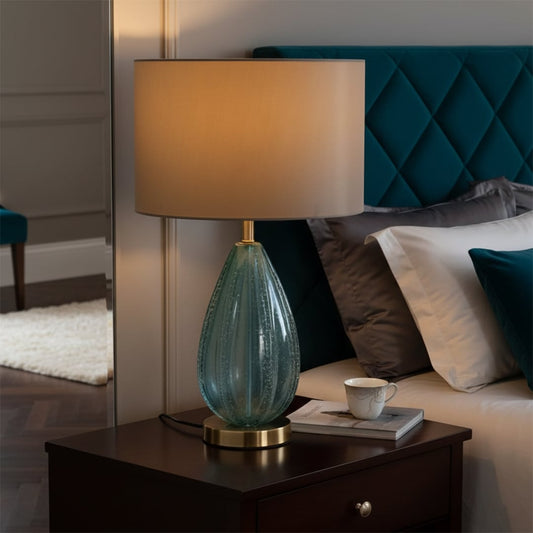 Teal glass table lamp with fabric shade placed beside a bed creating warm ambient lighting for modern bedrooms.