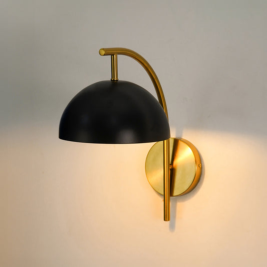 A modern wall-mounted sconce with a matte black dome shade and a brushed gold/brass curved arm and circular wall plate, shown lit against a plain light wall.