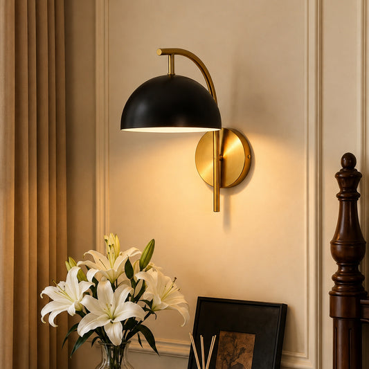 A modern wall-mounted sconce with a black dome shade and a brushed gold/bronze circular wall mount, shown lit in a warm beige room with curtains, white lilies, and a dark wooden post visible.