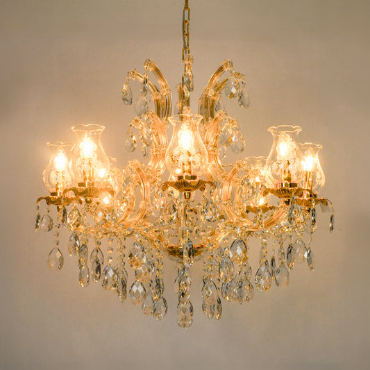 A multi-light crystal chandelier with a gold-tone metal frame and hanging clear glass teardrop prisms, shown illuminated against a plain beige wall.