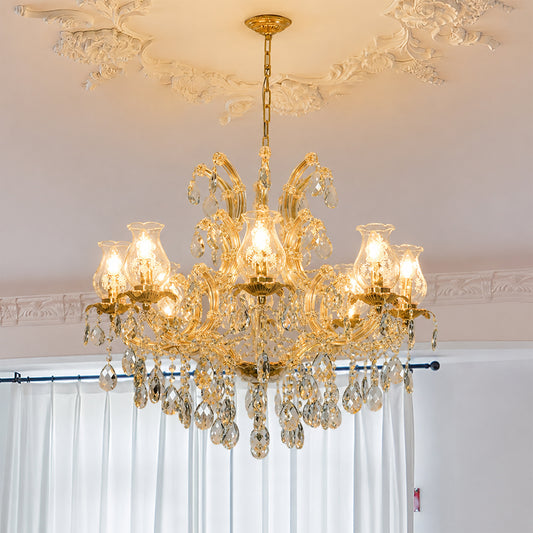 An ornate gold chandelier with multiple clear crystal hanging drops and bell-shaped glass shades, illuminated with warm light, hanging from a decorative ceiling in a room with white curtains.
