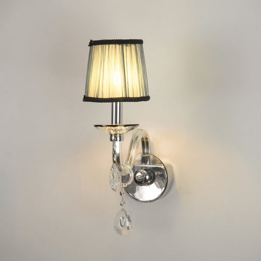 A wall-mounted sconce with a pleated beige fabric shade, black trim, a polished silver metal arm and wall mount, and clear hanging crystal teardrop ornaments, shown lit against a plain light wall.