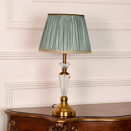 Decorative glass table lamp with gold metal base and pleated teal fabric shade placed on soft off white background.