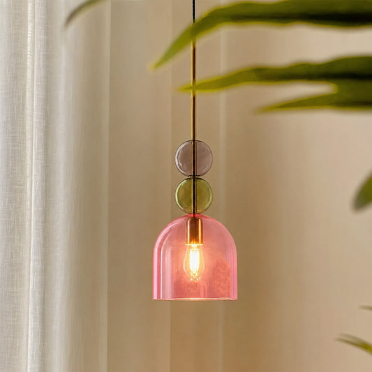 A modern pendant light with a translucent pink dome shade, a gold-toned metal rod, and three stacked circular glass accents in gray, green, and pink, shown hanging in a softly lit interior with curtain and green leaves in the background.