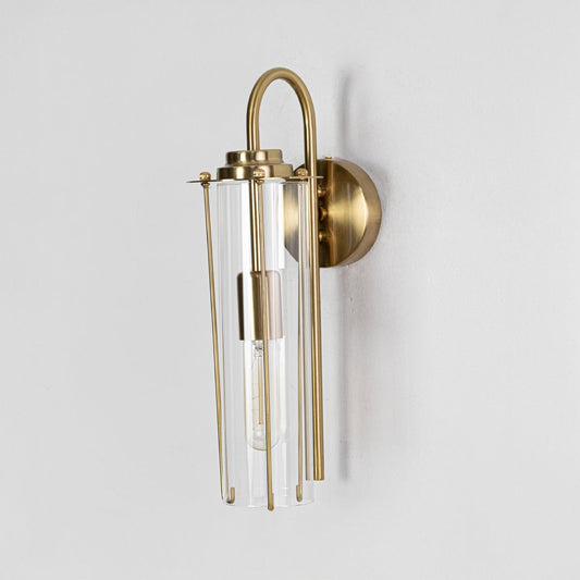A gold wall sconce with a tall cylindrical glass shade shown with the light off displaying its minimal contemporary structure in a clean setting.