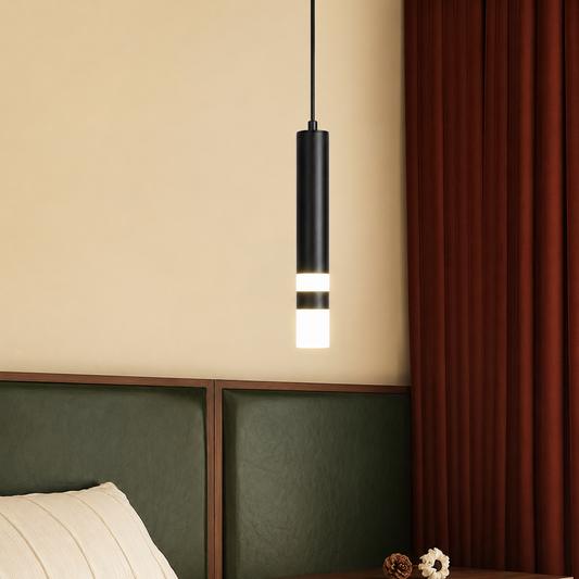 A modern cylindrical pendant light with a matte black upper body and glowing white lower section, hanging in a bedroom setting with a beige wall, dark green headboard, and rust red curtains.