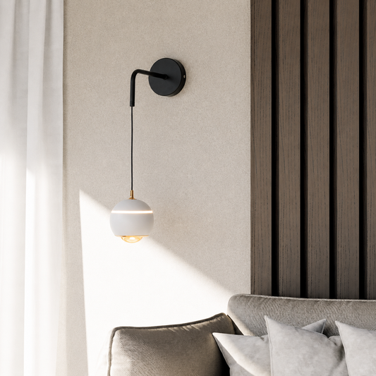 A modern wall-mounted sconce with a black metal arm and a white spherical shade, shown lit in a styled interior next to a sofa and textured wall paneling.