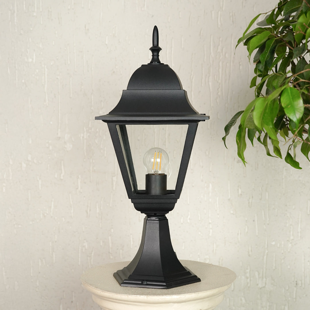Black outdoor gate light with warm glowing bulb styled beside plants for elegant exterior decor ambiance.