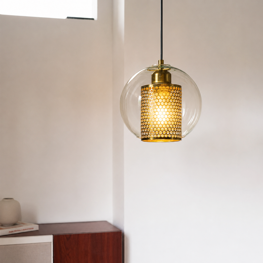 A modern pendant light with a clear glass globe shade, a brass-toned metal cap, and a honeycomb-patterned gold mesh diffuser, hanging in a minimalist room corner.
