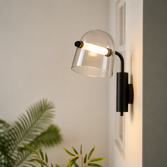 A modern wall-mounted sconce with a clear glass dome shade and a matte black metal body, shown lit against a light neutral wall with blurred green plants in the foreground.