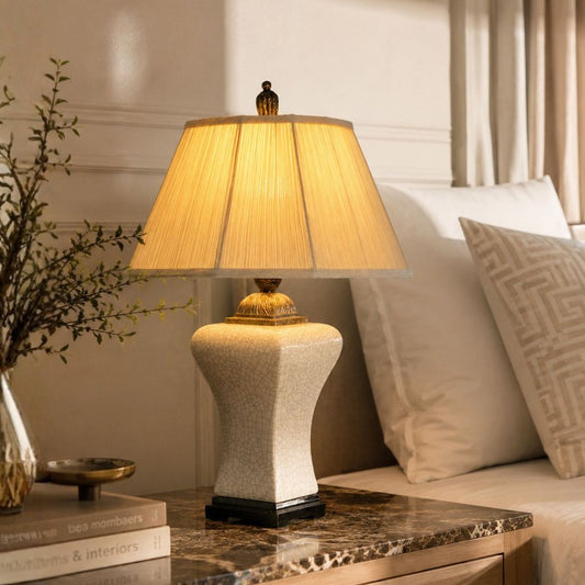 A table lamp with a textured cream-colored ceramic base and a pleated beige fabric shade, illuminated with a warm yellow glow. It is styled on a marble nightstand next to a sofa with pillows in a cozy bedroom setting.