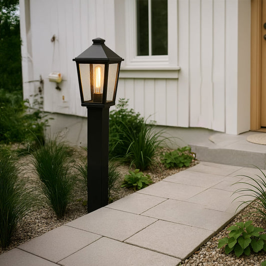 A modern outdoor pathway scene featuring a tall black post light turned on casting a warm glow beside a stone walkway near a white house entrance at dusk.