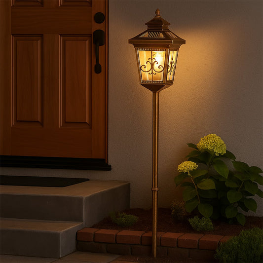 A bronze outdoor pathway stake light with the light on placed beside a front door casting a warm glow on the entry steps and garden area creating an inviting nighttime ambiance.