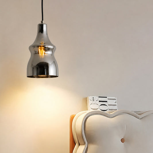 A modern pendant light with a polished chrome bell-shaped shade and a visible warm Edison bulb, hanging in a bedroom setting above a white tufted headboard against a beige wall.