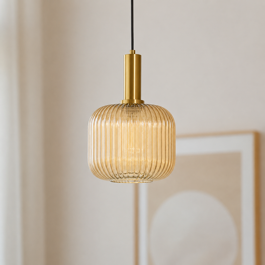 A modern pendant light with a ribbed, translucent amber glass shade and a brushed gold metal collar, suspended by a black cord against a warm beige wall.