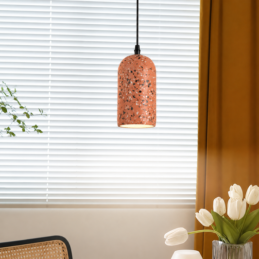 A terrazzo-patterned pendant light with a copper-orange shade hangs in front of white window blinds in a styled room setting with a vase of white tulips and a mustard curtain.