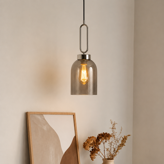 A modern pendant light with a smoky translucent glass shade and a brushed metal U-shaped hanger, shown hanging in a softly lit interior corner with a framed abstract print and dried flowers below.