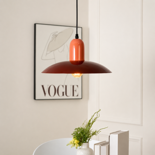 A modern pendant light with a glossy terracotta orange cylindrical shade and a wide matte dark red-brown metal dome shade, hanging in a styled interior setting with a framed VOGUE print on the wall.