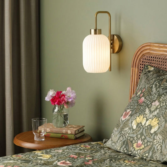 Gold metal wall light with white ribbed glass shade casting warm glow styled beside bed for cozy bedroom wall decor ambiance.