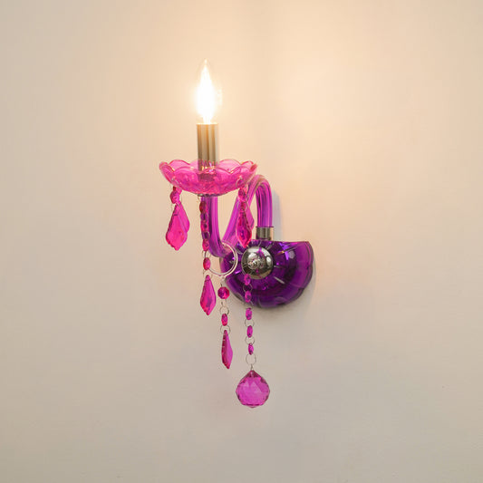 A decorative wall sconce with a lit candle-style bulb, featuring translucent pink and purple glass elements, hanging crystal drops, and a small silver-toned accent against a plain light wall.