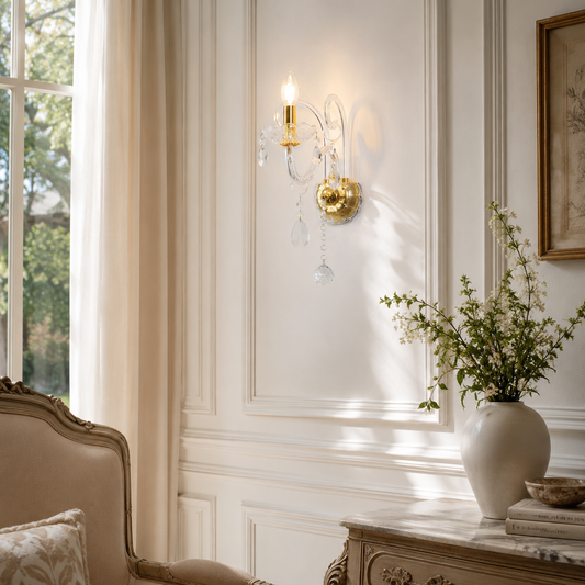 A gold wall sconce with clear crystal hanging drops is mounted on a white paneled wall in a bright, elegant room with a window, curtains, and a vase of greenery.