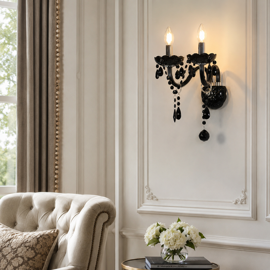 A black ornate wall sconce with candle-style bulbs and hanging crystal-like beads mounted on a white paneled wall in a styled interior with a tufted armchair and flowers.