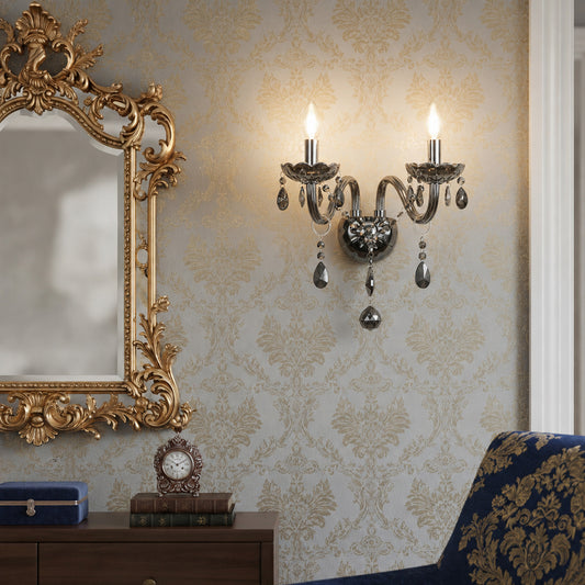 An ornate double-arm wall sconce with candle-style bulbs and hanging teardrop crystal drops, mounted on a damask-patterned wallpapered wall in a decorated room with a gold mirror and blue chair.