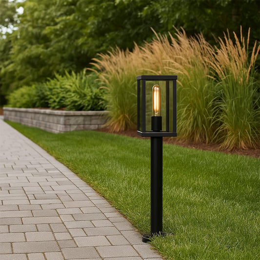 A black garden bollard light turned on beside a paved path casting a warm glow over green lawn and plants at dusk creating an inviting outdoor scene.