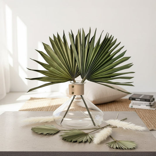 Natural green dried palm sun fan leaves arranged inside a clear glass vase on a grey table in front of a sunny window.