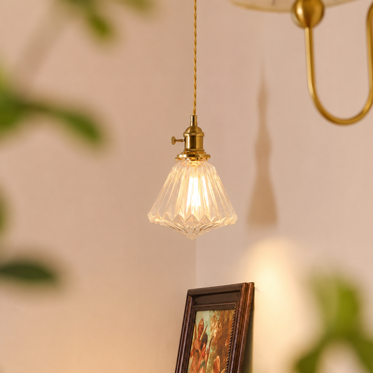 A vintage-style pendant light with a clear ribbed glass shade and a brass fixture hanging against a warm beige wall, with soft-focus decorative elements in the background.
