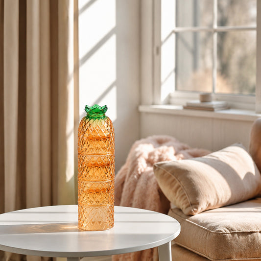 A translucent amber-orange pineapple-shaped glass vase with a green leafy top sits on a white round table in a sunlit living room next to a beige sofa.